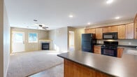 This single-floor home offers a thoughtfully designed open-concept layout that seamlessly connects the kitchen and living areas. The kitchen is outfitted with warm honey-toned wood cabinetry, dark countertops, stone-look tile flooring, and a full suite of black appliances including a refrigerator, range, built-in microwave, and dishwasher. A peninsula with additional counter space provides a natural divide between the kitchen and the living room. The living room is carpeted and bathed in natural light from a front-facing window, and features a ceiling fan and a tile-surround fireplace that serves as a focal point of the space. A front entry door with a large glass panel offers views of the neighborhood and allows additional light to filter in. The bedrooms are generously sized, each with plush carpeting, natural wood trim, and windows overlooking the surrounding neighborhood. One bedroom includes a spacious walk-in closet with wire shelving on multiple walls, offering ample storage. The bathroom conveniently incorporates a stacked washer and dryer unit alongside a full tub-shower combo, optimizing functionality within the space. Throughout the home, neutral wall tones, consistent wood trim, and recessed lighting create a clean, cohesive aesthetic.