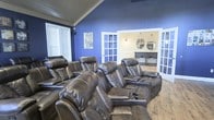 Located at 5303 Wilcox Rd in Dublin, Ohio, this single-floor property offers a thoughtfully designed living space with a standout feature: a dedicated home theater room that serves as the centerpiece of the home. The theater room is finished with bold cobalt blue walls and a warm wood-paneled vaulted ceiling that adds both height and character to the space. Multiple rows of plush, dark brown leather power recliner seats are arranged in a tiered layout, providing an immersive cinematic experience for a generous number of guests. Built-in cupholders and padded armrests add to the comfort and functionality of the seating arrangement. Natural light filters into the room through slatted window blinds, while elegant French doors with glass pane detailing connect the theater to an adjacent space, which appears to feature a vanity area with dual round mirrors and stylish wall sconces. Hardwood flooring runs throughout, adding a warm, cohesive feel to the interior. The overall ambiance of the home balances entertainment-focused design with comfortable, inviting finishes, making it a compelling option for those who value both style and function in a home setting.