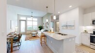 This thoughtfully designed one-bedroom, one-bathroom residence offers an efficient yet comfortable single-floor layout with a cohesive, modern aesthetic. The open-concept living, dining, and kitchen area forms the heart of the home, anchored by a striking granite-topped kitchen island with pendant lighting overhead. The kitchen features white cabinetry, stainless steel appliances, and light hardwood flooring that flows seamlessly throughout the entire unit. Large windows in the living area allow generous natural light to fill the space, offering outdoor views and creating an airy, welcoming ambiance. A dedicated dining nook provides an intimate setting for meals, complete with clean white walls that complement the overall neutral palette. The bedroom serves as a tranquil retreat, distinguished by a bold botanical-patterned accent wall and a large window framing outdoor green views. An adjacent walk-in closet adds practical storage. The full bathroom features granite countertops, white cabinetry, and a soaking tub with marble-style tile surround, maintaining the unit's clean, refined aesthetic. The consistent use of light hardwood flooring, soft neutral tones, and modern fixtures throughout creates a harmonious and inviting living environment.