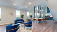 This well-appointed two-story space offers generously proportioned living across two nearly equal floors, connected by an elegant staircase with a rich wood banister and classic white balusters. The main floor is anchored by a dramatic open-plan living and dining area, distinguished by soaring double-height ceilings and floor-to-ceiling windows adorned with sheer draping curtains that flood the interior with natural light. A striking fireplace with a bold blue geometric accent wall serves as a focal point, complemented by warm hardwood flooring that flows throughout. The living area is thoughtfully arranged with plush navy blue velvet seating accented by gold-toned frames, centered on a soft area rug, while the adjacent dining space accommodates a large table with ample seating. The second floor mirrors the lower level's open, airy character, featuring its own lounge and seating areas bathed in natural light from multiple windows. Additional flexible lounging spaces are found across both floors, including a wall-mounted television area and intimate reading nooks with mid-century-inspired chairs. Recessed lighting, neutral walls, and consistent hardwood flooring throughout lend the entire space a cohesive, modern aesthetic that is both functional and inviting.