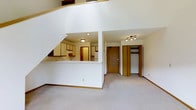 This two-story unit is designed with an open and functional layout that makes efficient use of its space across both levels. The main level features a carpeted living area that flows naturally toward the entry and a semi-open kitchen. The kitchen is defined by light-toned cabinetry, a pass-through counter that connects to the living space, and recessed and flush-mount ceiling lighting. A corridor leads to a closet with built-in shelving and hanging storage, as well as a door to the entry. Track lighting illuminates the hallway area, adding a practical touch. The staircase, visible from the main level, rises to the upper floor and features clean white walls that contribute to the bright, airy feel throughout the space. White walls and light carpeting are consistent design elements that create a cohesive, neutral interior. From the balcony, residents enjoy views of the surrounding grounds, which include manicured lawns, mature trees, winding walkways, and additional buildings within the community. The exterior of the multi-building community features brick construction with balconies and sloped rooflines, creating a classic residential aesthetic set within a landscaped environment.