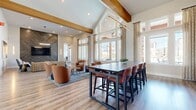 This thoughtfully designed single-floor community space offers generous square footage spread across a living room, a combined dining and living area, and a dedicated office. Throughout, light wood-tone flooring and crisp white walls create a clean, contemporary backdrop accented by bold teal and warm neutral tones.

The main living room is a striking focal point, featuring soaring vaulted ceilings with exposed wood beams and a dramatic wall of floor-to-ceiling windows that flood the space with natural light and provide views of the surrounding community. A herringbone-patterned wood accent wall frames a mounted television, while curated seating arrangements with warm terracotta lounge chairs invite comfortable gathering.

The combined dining and living area extends the social atmosphere, anchored by a long communal high-top dining table paired with upholstered bar-height chairs. A built-in wet bar with dark cabinetry, a textured tile backsplash, and open shelving lines one wall, complemented by bold teal accent walls adorned with curated artwork.

A secondary lounge area features a stunning marble-effect tile feature wall with a mounted television, vibrant blue accent chairs, and additional seating for more intimate gatherings.

The private office provides a quiet, functional workspace with a built-in desk running the length of the window, offering ample natural light and exterior views — ideal for focused work or study.