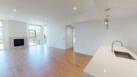 This single-floor space showcases a contemporary design characterized by clean lines, crisp white walls, and an abundance of natural light. The open-plan living area is anchored by warm hardwood flooring that extends throughout the main living zones, complemented by recessed lighting overhead. A floor-to-ceiling window wall floods the space with natural light and provides views of the surrounding greenery, while a stone-faced fireplace serves as a refined focal point.

The kitchen is outfitted with modern wood-grain cabinetry, white quartz countertops, stainless steel appliances including a French door refrigerator and gas range, and a geometric-patterned tile backsplash that adds visual interest. The layout flows seamlessly into the main living area, making it well-suited for everyday living and entertaining.

The bedrooms feature plush carpeting and mirrored sliding-door closets with built-in shelving and drawers. The primary bedroom benefits from direct outdoor access through glass doors. Two bathrooms serve the space: one features a frameless glass walk-in shower with white subway tile and a floating wood-toned vanity, while the second offers a tub-shower combination with large-format tile and a dual-sink vanity.