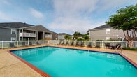 Located in Long Beach, Mississippi, this outdoor pool facility serves as a communal recreational amenity within a residential community. The centerpiece is a generously sized swimming pool with vivid turquoise water, framed by brick coping and a broad surrounding deck finished in aggregate concrete — a durable, slip-resistant surface well-suited to the Gulf Coast climate.

The pool deck is furnished with multiple lounge chairs and reclining seats, providing ample space for sunbathing and relaxation. A white vinyl perimeter fence encloses the pool area, clearly defining the space while maintaining an open, airy feel. Mature trees positioned just beyond the fence line offer a natural backdrop and occasional shade.

The facility includes a single-story pool house structure clad in gray horizontal lap siding with white trim — consistent with the architectural style of the surrounding multi-story residential buildings visible in the background. This structure houses separate men's and women's restroom facilities, each accessed via individual exterior doors along a covered concrete walkway. The building also features a secured entry door with a windowed panel.

Overall, the space presents a clean, functional, and inviting outdoor amenity designed to accommodate residents seeking leisure and recreation in a well-maintained community setting.