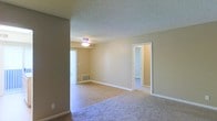 This single-floor residential space offers a straightforward and functional layout designed for comfortable everyday living. The main living area is a highlight of the space, featuring a combination of plush carpeting and hard surface flooring that clearly delineates different zones within the open floor plan. The walls are finished in a warm, neutral beige tone, creating a clean and inviting atmosphere throughout. A textured popcorn ceiling adds a classic character to the space, while a central ceiling-mounted light fixture with a decorative element provides ambient illumination to the living area. Natural light filters in through a large vertical-blind-covered window or sliding door, brightening the room during daytime hours. A floor vent indicates a built-in climate control system for year-round comfort. White baseboards and door trim offer a crisp contrast against the neutral walls. A doorway visible from the main living area leads to an adjoining room, suggesting a connected and practical layout. The overall ambiance is calm and understated, offering a blank canvas that is well-suited for personalization.