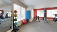 Located at 1870 Enterprise Drive in Virginia Beach, Virginia, this fitness center is a single-floor facility designed to support a comprehensive workout experience. The space is thoughtfully organized into distinct training zones, each catering to different aspects of physical fitness.

The cardio area features multiple treadmills and elliptical machines, positioned along bold red accent walls and flanked by large windows that invite natural light and outdoor views. Wall-mounted televisions provide entertainment during workouts. A ceiling fan and recessed lighting ensure comfortable conditions throughout.

The strength training section is equipped with a variety of selectorized cable and resistance machines, offering options for full-body conditioning. A dedicated free weights area includes a full rack of dumbbells, an adjustable weight bench, and a large floor-to-ceiling mirror that enhances the sense of space and supports proper form.

The functional training zone rounds out the facility with a collection of medicine balls, stability balls, exercise mats, and ab/back equipment, providing ample options for flexibility and core work. Neutral carpet flooring runs throughout, and large windows in every section ensure the space remains airy and well-lit. A water fountain and additional amenities are conveniently accessible near the facility's entrance.