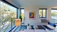 This thoughtfully designed single-floor space showcases a clean, contemporary aesthetic throughout. The living area is a standout, bathed in abundant natural light streaming through large, black-framed floor-to-ceiling windows that frame lush tree canopy views. Light hardwood flooring runs continuously through the space, contributing to an open and airy atmosphere. The galley-style kitchen is efficiently arranged and outfitted with stainless steel appliances including an electric range, over-the-range microwave, dishwasher, and refrigerator. Dark countertops contrast cleanly against crisp white cabinetry and a subtle gray subway tile backsplash, while open shelving adds functional display space. The bedroom offers a calm retreat with soaring ceilings, a ceiling fan with integrated lighting, and a large dark-paneled sliding closet door that adds a bold architectural element to the otherwise neutral palette. The bathroom is designed with a striking use of deep blue accent walls that frame a wide vanity with a white countertop, undermount sink, and chrome fixtures. A full-width mirror and wall-mounted lighting complete the polished look, while a window at the end of the hallway draws in natural light. The overall design balances modern minimalism with warm, livable touches throughout.