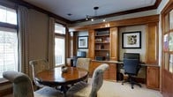 This single-story space exudes a warm, traditional aesthetic anchored by rich, dark-stained wood millwork and detailed crown molding throughout. A prominent built-in wall unit serves as the room's focal point, featuring open display shelving, closed cabinetry, and a granite-topped desk surface equipped with a desktop computer—ideal for business or personal use. Framed artwork is thoughtfully integrated into the built-in design, reinforcing a polished, curated atmosphere. Adjacent to the built-in, a separate workstation with a monitor provides additional functionality, making the space well-suited for productivity. A round wood dining or conference table surrounded by upholstered floral-patterned chairs occupies the opposite side of the room, offering a comfortable area for meetings or informal gatherings. Natural light filters through a large window dressed with textured drape panels, brightening the neutral-toned walls and light carpeted flooring. Overhead, a track lighting fixture supplements the natural light, ensuring the space remains well-lit throughout the day. The overall ambiance is sophisticated yet approachable, combining classic architectural details with practical amenities.