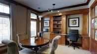 This single-story space exudes a warm, traditional aesthetic anchored by rich, dark-stained wood millwork and detailed crown molding throughout. A prominent built-in wall unit serves as the room's focal point, featuring open display shelving, closed cabinetry, and a granite-topped desk surface equipped with a desktop computer—ideal for business or personal use. Framed artwork is thoughtfully integrated into the built-in design, reinforcing a polished, curated atmosphere. Adjacent to the built-in, a separate workstation with a monitor provides additional functionality, making the space well-suited for productivity. A round wood dining or conference table surrounded by upholstered floral-patterned chairs occupies the opposite side of the room, offering a comfortable area for meetings or informal gatherings. Natural light filters through a large window dressed with textured drape panels, brightening the neutral-toned walls and light carpeted flooring. Overhead, a track lighting fixture supplements the natural light, ensuring the space remains well-lit throughout the day. The overall ambiance is sophisticated yet approachable, combining classic architectural details with practical amenities.