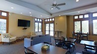 This single-floor communal space is thoughtfully designed to serve as both a relaxing lounge and a casual dining or gathering area. The room features a vaulted ceiling with a ceiling fan at its center, complemented by recessed lighting that ensures a well-lit environment throughout. Warm yellow walls contrast with the white ceiling and wood-trimmed windows, creating an inviting and comfortable atmosphere.

Large windows with wood framing line multiple walls, allowing generous natural light to fill the space while offering views of a surrounding outdoor area, including what appears to be a deck or balcony accessible through glass doors. The windows also feature transoms above, adding architectural interest and further enhancing the brightness of the room.

The lounge area is furnished with comfortable gray sofas and an accent chair with a patterned fabric, arranged around a central area. A wall-mounted flat-screen television serves as a focal point on one end. The dining section features multiple dark wood tables paired with matching chairs, arranged to accommodate small groups. Decorative greenery centerpieces add a natural touch to the tables, completing the welcoming and functional character of this shared space.