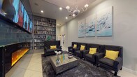 This single-floor lounge space offers a carefully curated interior that balances modern elegance with comfortable functionality. The room is anchored by a striking teal-tiled accent wall housing a wide linear electric fireplace with vivid amber flames, above which a row of flat-screen monitors is mounted for visual entertainment. Tall ceilings lend an airy, expansive quality to the space, further emphasized by the presence of a sculptural glass and chrome chandelier that serves as a refined focal point overhead.

Seating is arranged thoughtfully throughout, featuring charcoal tufted wingback chairs and a loveseat with nailhead trim detailing, all accented with vibrant yellow throw pillows that provide a bold contrast against the dark upholstery. A textured stone-finish coffee table sits atop a plush, deep-pile charcoal area rug, grounding the seating arrangement over light-toned tile flooring.

One wall features floor-to-ceiling built-in shelving styled with books, decorative objects, and a globe, adding depth and character to the room. On the opposite side, a triptych of abstract aquatic-themed artwork in soft blues and whites adorns the neutral gray walls, contributing to the overall polished and contemporary ambiance. Sheer curtains filter natural light near the windows, completing the refined atmosphere.
