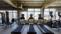 Located at 3946 Saint Johns Ave in Jacksonville, Florida, this fitness center offers a well-equipped, single-floor workout facility designed with both functionality and aesthetics in mind. The space features an industrial-chic aesthetic, with exposed ceiling ductwork and piping set against clean white walls and smooth wood-look flooring that runs throughout. Large floor-to-ceiling windows line one side of the space, flooding the interior with natural light and offering scenic views of palm trees and the surrounding landscape — an energizing backdrop for any workout. The cardio zone is anchored by a row of Matrix treadmills positioned directly in front of the windows, allowing users to enjoy the outdoor scenery while exercising. Additional cardio equipment, including elliptical machines, is arranged along the same wall. A dedicated strength training area features a multi-functional cable and resistance rig, a heavy punching bag, TRX suspension trainers, medicine balls, kettlebells, and an adjustable bench, providing a comprehensive range of training options. Wall-mounted televisions are positioned throughout the space for entertainment. A staircase with black metal railings is visible, and mirrored walls in the strength zone enhance the sense of openness. The overall ambiance is bright, modern, and motivating.