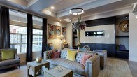 This single-floor space is designed with a refined, contemporary aesthetic that blends comfort and sophistication. The centerpiece of the lounge area is a pair of tufted leather sofas adorned with colorful accent pillows in muted rose, yellow, and sage tones, arranged around wooden coffee tables that display decorative sculptural pieces. A striking ring-shaped pendant light hangs from a tray ceiling, casting a warm, ambient glow throughout the room. Large windows dressed with floor-to-ceiling dark drapes allow natural light to filter in while framing views of the surrounding exterior. A trio of vibrant abstract artworks mounted on a warm-toned accent wall adds a bold visual focal point, complemented by a sputnik-style chandelier nearby. Toward the rear, a white subway tile feature wall with a dark inset niche provides a clean, contrasting backdrop, hinting at an adjacent bar or service area. A tall brass vase with an oversized floral arrangement adds dramatic vertical interest. The overall ambiance is polished and inviting, with layered lighting, rich textures, and curated décor creating a well-appointed communal environment.