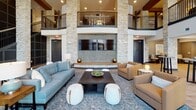 This impressive communal space is defined by its dramatic double-height ceilings and striking stacked stone columns that anchor the design and draw the eye upward toward an open upper-level mezzanine with sleek metal railings. The grand lobby lounge area serves as the heart of the space, featuring a large flat-screen display mounted against a textured stone feature wall, surrounded by carefully arranged seating including a tufted blue sofa, neutral armchairs, and rounded ottomans atop a patterned area rug — creating an inviting and social atmosphere.

Expansive floor-to-ceiling windows line one side of the space, framed by sheer linen drapes that soften the abundant natural light while offering views of the surrounding exterior courtyard. The warm hardwood flooring runs throughout, complementing the rich blend of natural stone and dark architectural millwork.

A fully appointed kitchen area is also part of this space, outfitted with dark cabinetry, a large curved kitchen island with seating, stainless steel appliances including a range and refrigerator, a white subway tile backsplash, and pendant lighting — ideal for communal gatherings or events. A staircase with dark metal railings connects to the upper mezzanine level, adding to the layered, multi-dimensional character of this well-appointed space.