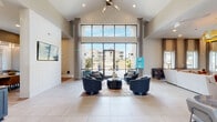 This single-story residential clubhouse is designed to foster community with a sophisticated blend of modern aesthetics and functional gathering spaces. The grand entrance lobby immediately impresses with soaring vaulted ceilings, large-format light tile flooring, and a dramatic floor-to-ceiling glass curtain wall that floods the space with natural light and offers unobstructed views of the surrounding community. A curated seating arrangement of plush navy swivel chairs anchors the welcoming lounge area beneath a sleek ceiling fan fixture.

Moving through the space, a comfortable media lounge features rich warm-toned furnishings, a large wall-mounted television set into a textured dark wood accent wall, and a bold gold sculptural chandelier that adds a striking design element. Adjacent to this area, a fully equipped wet bar with a marble-topped island, leather bar stools with gold ring accents, pendant lighting, and a tiled backsplash provides an ideal social hub.

A dedicated co-working area offers individual workstations, soft lounge seating, and tall windows that invite natural light, while a dining nook with a solid wood table and leather chairs provides a versatile gathering spot. Throughout, elevated ceilings, layered lighting, and thoughtful material selections create a polished yet inviting ambiance.