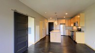 This single-floor space showcases a thoughtfully designed open-concept layout that seamlessly blends functionality with modern style. The kitchen serves as the heart of the space, featuring light maple cabinetry, a center island with clean-lined cabinetry below, and a suite of stainless steel appliances including a refrigerator and gas range. Pendant lighting above the island adds warmth and visual interest, complemented by recessed ceiling lights that illuminate the area evenly throughout. The dark hardwood flooring runs continuously across the open living and kitchen areas, providing a rich contrast to the neutral-toned walls and light cabinetry. A standout architectural feature is the sliding barn door, finished in a deep charcoal tone, which adds a contemporary industrial touch while offering a space-saving room divider solution. The corridor beyond leads to additional rooms, hinting at a well-organized floor plan. The smooth, neutral-painted walls and high ceilings contribute to an airy, spacious feel throughout. Overall, the space strikes a clean, modern aesthetic with practical design choices that make it both livable and visually appealing.
