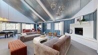 Located at 3205 W Floyd Ave in Denver, CO, this thoughtfully designed single-floor community space offers a diverse mix of functional and social areas across a generously sized footprint. The centerpiece is a dramatic main lounge distinguished by soaring vaulted ceilings clad in corrugated metal panels, lending an industrial-chic aesthetic that is both bold and inviting. Deep slate-blue board-and-batten walls, globe chandelier pendants, and a classic white fireplace anchor the room, while plush velvet sofas, mid-century accent chairs in warm terracotta, and a built-in shelving unit create a layered, curated atmosphere. Natural light filters through curtained windows throughout. A separate meeting or dining nook features a large picture window with outdoor tree views, a built-in concrete-style shelf, and a round table with modern seating — ideal for small gatherings or focused work. A spacious game room with neutral walls and soft carpet provides room for shuffleboard and foosball, offering a bright, casual retreat. An additional flexible room serves as a fitness or activity studio, equipped with full-length mirrors, a wall-mounted screen, and an open carpeted floor. A transitional lounge area with warm wood flooring and paired bistro tables rounds out the space, connected seamlessly via sliding barn doors for versatile use.