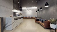 This contemporary two-floor commercial space makes an immediate impression with its grand lobby, where a sculptural marble reception desk anchors the entrance beneath cascading pendant lighting. Floor-to-ceiling steel-framed glass doors flood the entryway with natural light, while polished concrete floors extend throughout, providing a clean, industrial foundation that complements the refined finishes above.

The lobby features a geometric white tile accent wall with an intricate diamond pattern, serving as a dramatic backdrop to a built-in wooden bench lined with navy cushions. Nearby, a curated lounge area showcases rich cognac leather sofas, modern accent chairs, and dark textured tile walls that add warmth and depth to the space.

A wood-paneled elevator bank and stairwell corridor offer practical connectivity between floors. The double-height co-working area on the main floor is a standout, featuring long walnut communal work tables, ergonomic seating, and a mezzanine level accessible via a bold black steel staircase with open railings. The upper level hosts additional lounge and seating zones with deep navy sofas, warm floor lamps, and bar-height tables. Throughout both floors, carefully layered lighting, quality materials, and a cohesive dark-toned palette create a professional yet inviting atmosphere.