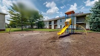 Located at 1900 Cardinal Trail Dr in Galloway, Ohio, this outdoor community space serves as a shared recreational area within a residential complex. The space is defined by an open, single-level layout that provides ample room for outdoor activity and leisure.

At the heart of the area is a playground structure featuring a prominent yellow slide and climbing tower with a covered roof, supported by a sturdy blue metal frame. The play area is surfaced with wood chip mulch, providing a safer ground cover for children at play. Adjacent to the main playground is an additional play element, visible in the background, extending the recreational options available.

The surrounding grounds are landscaped with open grassy areas and mature trees that provide natural shade and greenery, contributing to a welcoming outdoor atmosphere. The residential buildings that border this space are two-story structures with light gray vinyl siding, red accent doors, and private balconies, creating an enclosed, community-oriented setting.

The space is well-suited for residents seeking an active outdoor environment, particularly for families with children. The combination of open lawn space and dedicated play equipment makes this area a functional and practical community amenity within the Galloway neighborhood of Columbus, Ohio.