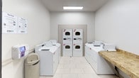 This shared Laundry Center is a well-organized, single-floor facility designed to meet the everyday needs of its users. The space is finished with light-toned large-format tile flooring and neutral walls, creating a bright and clean environment throughout.

The main laundry area is equipped with multiple top-load washing machines and both stacked and standalone dryers, providing ample capacity for simultaneous use. A granite-topped folding counter offers a practical surface for sorting and folding laundry. Machines are clearly numbered and serviced through CSC, with posted service instructions for easy reference.

An adjacent entry area connects the laundry room to the rest of the facility and features a distinctive Luxer One smart package locker system — a secure and convenient solution for parcel delivery and pickup. A horizontal rail runs along the lower portion of the walls in this transitional space, adding a subtle architectural detail while offering functional protection.

Overhead flush-panel lighting ensures the entire facility is well-lit, and a security camera is visible, providing an added layer of safety. The overall ambiance is utilitarian yet tidy, reflecting a thoughtfully maintained communal space built for practicality and ease of use.