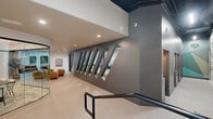 This thoughtfully designed single-floor commercial space offers a dynamic blend of contemporary architecture and curated interior design. The layout encompasses open lounge areas, well-appointed hallways, a formal conference/dining room, and several versatile rooms connected by wide, light-toned tile corridors.

A standout architectural feature is the dramatic interior wall fitted with angled, diamond-shaped windows that peer into adjacent spaces, creating visual connectivity and depth throughout the floor. Black-framed glass partitions and doors add an industrial-modern contrast to the crisp white walls.

The open lounge area is anchored by a rich terracotta velvet sofa, a low-profile wooden coffee table, and deep-green arched booth seating tucked into accent-painted alcoves — all set beneath a striking exposed black ceiling with visible ductwork and linear lighting. A separate seating nook features rounded, caramel-toned chairs arranged around a marble-top accent table.

The conference room is enclosed by full-height glass walls and features a long wooden table with upholstered boucle chairs, patterned area rugs, curated artwork, and a tall potted olive tree. Throughout, the space maintains a cohesive, upscale aesthetic with polished tile flooring, recessed lighting, and carefully selected furnishings that balance function with refined style.