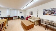 Located at 25 Lorne Ave in Newmarket, Ontario, this single-floor combined living and dining space offers a functional and communal layout suited for group gatherings and everyday interaction. The interior features light hardwood-style flooring throughout, contributing to a clean and cohesive look across both areas.

The living area is generously furnished with a large beige sofa, a substantial rectangular coffee table, and additional seating including armchairs in varying styles. A wall-mounted decorative mirror and framed artwork add character to the neutral white walls, while large vertical blinds allow natural light to filter in from the windows.

The dining area accommodates multiple dining tables — including a rectangular table with several chairs and a round table — making it well-suited for communal dining. The walls are adorned with a world map, framed photos, and artwork, giving the space a personalized, lived-in feel. A bulletin board near the entrance displays notices and community information, reinforcing the space's communal purpose.

Overhead fluorescent lighting provides consistent illumination throughout. A wood-toned door with an elevator sign nearby indicates convenient accessibility. Overall, the space presents a practical and welcoming environment, thoughtfully arranged to support both social and daily living activities.