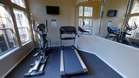 This single-story fitness center in Arlington, Texas, offers a focused and functional workout environment designed to meet essential exercise needs. The space is finished with neutral beige walls and dark carpeted flooring, creating a practical and clean atmosphere conducive to exercise. Natural light enters through a window, brightening the room and providing views of the surrounding greenery outside, adding a pleasant outdoor connection to the interior space. A large wall-mounted mirror runs along one side of the room, visually expanding the area and allowing users to monitor their form during workouts. The fitness center is equipped with key cardio machines, including a treadmill and an elliptical trainer, positioned to make efficient use of the available floor space. A wall-mounted flat-screen television is installed at a comfortable viewing height, allowing users to watch programming during their workout sessions. Posted fitness center rules ensure the space remains organized and accessible for all users. Overall, the space delivers a straightforward, well-maintained fitness environment suited for daily cardio routines.
