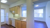 This single-floor residence offers a clean, contemporary layout designed for comfortable everyday living. The kitchen is a standout feature, outfitted with light maple cabinetry that extends from base to upper levels, providing ample storage space. A double-basin sink sits beneath a window, allowing natural light to pour in while completing everyday tasks. Light countertops complement the warm wood tones, and a white dishwasher is seamlessly integrated into the cabinetry for a cohesive look. Recessed lighting overhead keeps the kitchen well-illuminated at any hour. A structural column subtly defines the transition between the kitchen and the adjoining living area, maintaining an open and connected feel without fully dividing the spaces. The living area is bright and airy, benefiting from large windows that frame views of the surrounding outdoor scenery, inviting an abundance of natural light. Rich hardwood-style flooring grounds the living space, adding warmth and visual depth. The overall ambiance of the residence is fresh and inviting, with a neutral palette that provides a versatile backdrop, making it easy to personalize and style to any taste.