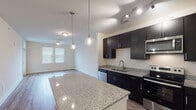 This single-floor residence offers a thoughtfully designed layout that balances functionality with contemporary style. The kitchen serves as a central highlight, showcasing dark espresso cabinetry that extends to the ceiling for ample storage, granite countertops, and a full suite of stainless steel appliances including a refrigerator, electric range, built-in microwave, and dishwasher. A spacious island with matching granite provides additional workspace and casual seating. Pendant lighting with globe fixtures hangs above the island, while track lighting illuminates the kitchen workspace. The kitchen opens directly to the living area, which features warm wood-look laminate flooring and a ceiling fan, creating a cohesive open-concept flow. Natural light enters through blinded windows. A carpeted bedroom offers a ceiling fan and well-sized windows that bring in natural light, while a secondary room features the same wood-look flooring and windows. The bathroom includes dark vanity cabinetry with granite countertop, a large mirror, and access to a walk-in closet with built-in shelving. Consistent finishes and neutral tones throughout contribute to a clean, modern aesthetic.