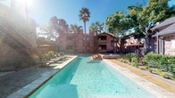 This single-floor residence is set within a charming multi-story brick building, surrounded by mature palm trees and lush greenery that create a warm, resort-like atmosphere. The community features a generously sized outdoor pool flanked by lounge seating and shaded areas, offering a serene retreat for relaxation.

Inside, the unit presents a clean, contemporary aesthetic with neutral white walls and dark stained concrete flooring that adds an industrial-chic character throughout. The dining area is a notable highlight, anchored by a striking round marble-top table paired with rattan chairs that blend elegance with casual comfort. A large, framed abstract artwork adorns the wall, adding a sophisticated artistic touch to the space.

A sliding barn door provides a functional and stylish transition between rooms, reinforcing the modern design sensibility found throughout. Recessed lighting and well-placed ceiling vents contribute to a comfortable and well-lit environment. The overall ambiance is one of understated refinement — a thoughtfully curated space where natural materials, artistic accents, and clean architectural lines come together cohesively to create a welcoming and visually engaging living environment.