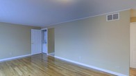 This single-floor space presents a clean, straightforward layout with classic architectural details. The main living area is finished with warm hardwood flooring that runs throughout, lending a timeless quality to the interior. Walls are painted in a soft, neutral beige tone, complemented by crisp white baseboards and crown molding that frame the room with a polished look. The smooth, light-colored ceiling contributes to a bright and open atmosphere.

A notable feature of this space is the in-unit laundry, visible through an open doorway. A stacked washer and dryer unit is neatly tucked into a dedicated alcove or closet, offering a practical and convenient amenity within the unit itself. The six-panel white interior door adds a traditional design touch and provides clear separation between the living area and the laundry space.

The overall ambiance of the space is understated and move-in ready, with a neutral palette that provides a versatile backdrop for a variety of furnishing styles. The hardwood floors and classic trim details give the space a sense of quality and durability, while the open layout allows for flexible furniture arrangement.