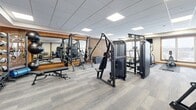 This 820 square foot fitness center in Savage, MN, offers a comprehensive workout experience in a single-floor layout. The space is characterized by its modern design, featuring a harmonious blend of functionality and aesthetics. The center boasts a variety of exercise equipment, catering to both cardio and strength training needs. Large windows allow natural light to flood the area, creating an energizing atmosphere for workouts.

The interior is adorned with a combination of white walls and warm wood accents, particularly noticeable in the vertical slat wall treatments. These elements contribute to a welcoming and contemporary ambiance. The flooring consists of durable, low-maintenance gray carpet tiles, ideal for a fitness environment.

The gym is equipped with state-of-the-art machines, including treadmills, ellipticals, and stationary bikes in the cardio section. The strength training area features weight machines, free weights, and a multi-functional cable machine. Additional amenities include a water station and strategically placed mirrors to assist with proper form during exercises.

Ceiling-mounted LED panels provide even, bright lighting throughout the space. The layout is open and spacious, allowing for easy navigation between different workout zones. This well-designed fitness center offers a comfortable and efficient environment for users to pursue their health and wellness goals.