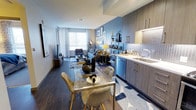 Unit 702 is a thoughtfully designed 1-bedroom residence spread across a single floor, offering a seamless open-concept layout that connects the kitchen, dining, and living areas into one cohesive living space. The kitchen is outfitted with sleek, wood-toned cabinetry featuring gold hardware, a subway tile backsplash, stainless steel appliances including a dishwasher, and a deep double-basin sink — combining both style and functionality. A glass-top dining table with modern gold-accented chairs sits at the heart of the space, providing an elegant transition between the kitchen and the living area. The living room is anchored by a bold blue accent wall adorned with curated wall art, creating a vibrant focal point. Rich, dark hardwood-style flooring runs throughout, complementing the warm neutral tones of the furnishings. A large window floods the space with abundant natural light, enhancing the open and airy ambiance. Recessed lighting and carefully placed accent lamps add warmth and depth. The overall aesthetic is contemporary and curated, blending modern finishes with inviting design elements that make this residence both functional and visually striking.