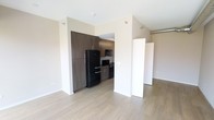 This single-floor space combines modern design with functional living. The open-concept layout creates a seamless flow between the living area and the kitchen, making the space feel airy and well-connected. The kitchen is outfitted with rich, dark wood cabinetry that extends floor to ceiling, providing ample storage. Black appliances — including a full-size refrigerator, microwave, and range — lend a sleek, contemporary edge to the culinary space. A subtle tile backsplash adds texture and visual interest. The living area is finished with light-toned hardwood-style flooring that runs throughout, brightening the interior and offering a clean, cohesive aesthetic. Neutral warm-white walls allow natural and ambient light to reflect evenly across the space. Architectural details such as an exposed ventilation duct add an industrial character, complementing the modern design sensibility. A structural column with a clean white finish serves as a design element that subtly defines the transition between the kitchen and living zones. The overall ambiance is contemporary and understated, offering a well-balanced foundation for a variety of interior styles.