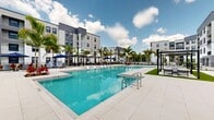 Located on Stockton Street in North Fort Myers, Florida, this outdoor amenity space showcases a resort-style pool area designed for relaxation and recreation under the Florida sun. The centerpiece is a generously sized swimming pool with crystal-clear turquoise water, varying in depth and offering wide, gently sloped entries for easy access. The surrounding deck is finished in light-toned large-format tile, providing a clean, contemporary aesthetic throughout. Rows of sleek lounge chairs line the perimeter, complemented by shaded cabana-style seating areas equipped with modern pergolas and canopies outfitted with ceiling fans, offering comfortable retreats from the sun. Vibrant tropical landscaping, including tall swaying palm trees, colorful accent plantings, and manicured garden beds bordered by black metal fencing, enhances the resort atmosphere. A dedicated turf lounge island with additional seating provides a lush gathering spot within the deck area. Blue market umbrellas are strategically positioned throughout, adding pops of color against the neutral hardscape. The entire space is framed by contemporary multi-story residential buildings with modern facades, balconies, and large windows, creating an enclosed, community-oriented environment ideal for outdoor leisure.
