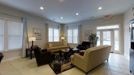 This single-floor space centers around a spacious, thoughtfully arranged living room that offers a comfortable and functional layout. The room is illuminated by a series of recessed ceiling lights, providing consistent, even lighting throughout. Multiple windows fitted with white plantation shutters and sheer curtains allow natural light to filter in, enhancing the room's bright and airy atmosphere. A glass-paneled entry door on one side further contributes to the natural light and connects the interior to the outdoors.

The living room is furnished with a coordinated seating ensemble, including a beige sofa, two dark upholstered armchairs, and a matching loveseat, all arranged around a dark wood coffee table. A patterned area rug anchors the seating area, adding texture and visual interest to the light-toned tile flooring. Framed wall art and a tall potted plant add decorative accents, while floor lamps supplement the ambient lighting. The neutral color palette of beige, brown, and cream tones creates a cohesive and inviting environment throughout the space.