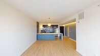 This modern single-floor residence showcases a thoughtfully designed open-concept layout that seamlessly integrates the kitchen and living areas. Light hardwood flooring flows throughout the main living space, creating a warm and cohesive aesthetic. The kitchen is a standout feature, centered around a striking blue island with a light countertop that serves as both a workspace and gathering point. Dark-toned cabinetry reaches toward the ceiling, complemented by a tiled backsplash and a full suite of stainless steel appliances, including a refrigerator, electric range, and built-in microwave. Globe pendant lights add a stylish finishing touch overhead. The living area is open and airy, bathed in natural light from large sliding glass doors that open to an outdoor view. A spacious bedroom features plush carpeting, a ceiling fan, and a window framing views of the surrounding neighborhood. The bathroom is clean and contemporary, with wood-toned cabinetry, a white countertop, a large mirror with wall sconces, and a full bathtub. The entry corridor provides a practical transition into the home, with the kitchen visible just beyond the front door. Subtle accent lighting along the hallway adds warmth and depth to the overall ambiance of this well-appointed residence.