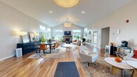Located at 1 Juniper Dr in Delmar, NY, this single-floor space is defined by its open, airy layout and thoughtful architectural design. Soaring vaulted ceilings rise dramatically throughout the main living area, creating an impressive sense of volume and grandeur. Rich hardwood flooring runs continuously through the space, lending warmth and cohesion to every zone.

The main gathering area is anchored by a striking stone fireplace with a mounted television above, serving as a natural focal point. Multiple seating arrangements — including plush sofas, swivel accent chairs, and side seating — offer flexible configurations suited to a variety of uses. Large windows line the walls on multiple sides, inviting abundant natural light and offering pleasant outdoor views of the surrounding greenery.

A dedicated dining area features a sleek table and leather chairs alongside a dark credenza, positioned adjacent to additional windows for a bright, welcoming atmosphere. The entry foyer is equally impressive, featuring soaring ceilings, an octagonal skylight, and glass double doors that frame natural views from the moment of arrival.

Additional rooms visible from the main area provide functional utility space. Throughout, the palette remains neutral and clean — warm whites, soft grays, and natural wood tones — creating an inviting and versatile environment. The seamless flow between areas and the abundance of natural light make this a bright, well-designed, and highly functional space.