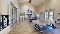 This expansive 820 square foot recreation room is a testament to modern design and functionality. The space boasts impressively high ceilings with exposed wooden beams, creating an open and airy atmosphere. The room is illuminated by a combination of recessed lighting and decorative chandeliers, complementing the abundance of natural light that floods in through multiple sets of French doors.

The flooring consists of high-quality wood-look planks, providing durability and a warm aesthetic. The room is equipped with an array of entertainment options, including multiple game tables such as air hockey, pool, and ping pong, catering to diverse recreational preferences. A sleek, modern fireplace with a stylish tiled surround serves as a focal point, adding both warmth and visual interest to the space.

Wall-mounted TVs and tasteful artwork adorn the crisp white walls, enhancing the room's modern appeal. Strategically placed potted plants bring touches of greenery, softening the overall look. The room's layout is thoughtfully designed to accommodate group activities while maintaining a sense of spaciousness.

This versatile space seamlessly blends comfort and entertainment, making it an ideal setting for social gatherings or relaxed leisure time. The attention to detail in both design and amenities creates an inviting atmosphere that caters to a variety of recreational needs.