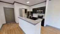 This single-floor residence offers a clean, contemporary design with thoughtful finishes throughout. The kitchen serves as the focal point of the space, showcasing a large peninsula with striking granite countertops and an integrated sink, providing ample prep space and a natural gathering area. Rich, dark cabinetry with raised panel doors creates a bold contrast against the white subway tile backsplash and crisp white walls. Stainless steel appliances, including a refrigerator, range, and over-the-range microwave, complete the modern kitchen aesthetic. Light hardwood-style flooring flows throughout, lending warmth and continuity to the open layout. Crown molding along the ceiling adds a refined architectural detail, elevating the overall finish level of the space. A doorway visible from the kitchen hints at additional rooms beyond, suggesting a functional and well-organized floor plan. Recessed lighting ensures the space is well-illuminated, enhancing the bright and welcoming atmosphere. Overall, the residence balances practical design with contemporary style, making it an inviting and well-appointed living environment.