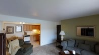 This single-floor residence offers a thoughtfully arranged layout that balances comfort and functionality. The living room serves as the heart of the home, featuring plush carpeted flooring, neutral white walls accented by a warm olive-green feature wall, and ample space for relaxed seating arrangements. A classic armchair with decorative cushions and a dark wood side table with a traditional table lamp add a timeless, homey character to the space. A dark wood coffee table anchors the seating area, completing the inviting atmosphere.

The kitchen is accessible directly from the living area through an open passageway, creating a seamless flow between the two spaces. It features warm wood-toned cabinetry, a white dishwasher, a double sink with under-cabinet lighting, and light-colored countertops that provide a practical and welcoming cooking environment. The kitchen's tile or laminate flooring contrasts with the carpeted living area, clearly defining each functional zone.

Overall, the space presents a clean, well-maintained interior with a warm and unpretentious aesthetic. The open connection between the living and kitchen areas promotes a sense of spaciousness, making this residence a comfortable and practical place to call home.