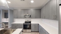 This single-floor residence offers a functional and modern layout across three bedrooms and three full bathrooms, making it well-suited for a variety of living arrangements. The entrance opens into a bright, open-concept kitchen and living area finished with light wood-look flooring. The kitchen is a standout feature, showcasing grey shaker-style cabinetry extending to the ceiling for abundant storage, crisp white countertops, stainless steel appliances including a range and over-the-range microwave, and a central island with pendant lighting that doubles as a casual dining and prep space. The living area benefits from natural light through large windows dressed with white blinds and is accented by a ceiling fan overhead. The three bedrooms are carpeted for comfort, each equipped with a ceiling fan and window for natural light. The primary bedroom is a generous size with its own dedicated closet featuring built-in shelving and hanging rods. Each bathroom offers clean, contemporary finishes with white countertops and framed vanity mirrors. A dedicated hallway connects the various rooms, contributing to a logical and organized flow throughout the home. Two closets provide additional storage. The overall aesthetic is modern and neutral, with grey tones, clean lines, and consistent finishes throughout.