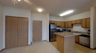 This single-floor space features an open-concept kitchen that serves as a central highlight of the layout. The kitchen is outfitted with warm oak cabinetry providing generous storage, complemented by granite-style countertops that add a polished, durable surface for meal preparation. Stainless steel appliances, including a full-size refrigerator, electric range, and microwave, lend a clean, modern aesthetic to the space.

A peninsula counter extends from the main kitchen area, creating a natural divide between the kitchen and the adjacent living space while also offering additional counter space. Overhead fluorescent lighting ensures the kitchen is well-illuminated throughout.

The entryway is conveniently positioned near the kitchen, featuring two solid wood doors — likely a unit entry and a closet — with light wood finishes that complement the overall warm tone of the interior. The flooring transitions from light hardwood in the kitchen area to a neutral carpet elsewhere, adding subtle texture variation throughout. The neutral wall tones and practical finishes give the space a clean, functional ambiance suited for everyday living.