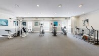 Located at 12400 Castlebridge Dr in Jersey Village, Texas, this fitness center offers a spacious, single-floor exercise room designed to support a well-rounded workout routine. The room is finished with neutral-toned carpeting and two-tone walls featuring a crisp white upper half and a soft grey wainscoting below, giving the space a clean, polished appearance. Recessed lighting throughout ensures even illumination across the entire floor area, while multiple windows along one wall invite natural light and create an open, airy atmosphere.

The equipment layout is thoughtfully organized to maximize the available floor space. Cardio offerings include elliptical trainers, a stationary bike, and a treadmill, while strength training is supported by a multi-function cable machine and additional resistance equipment. Decorative artwork with nature-inspired and animal motifs is displayed along the walls, adding a lively visual element to the environment.

Structural columns with white trim accents define distinct zones within the room, and tile flooring at the entry transitions smoothly into the carpeted workout area. A potted plant near the exit adds a touch of greenery. This well-maintained fitness facility provides residents with convenient access to a comprehensive range of exercise options in a comfortable and welcoming setting.