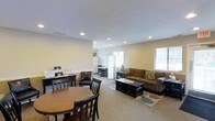 This single-floor space is designed with an open-concept layout that seamlessly blends a dining area and a living room into one cohesive environment. The dining area is furnished with a warm-toned round wooden table and matching dark chairs, positioned near a functional desk or sideboard along the wall — ideal for both casual meals and everyday tasks. Adjacent to it, the living area features a plush brown sofa accented with decorative pillows, centered around a dark wooden coffee table set atop a patterned area rug, adding texture and definition to the carpeted floor. Recessed lighting embedded in the white ceiling provides consistent, even illumination throughout the space, while multiple windows dressed with horizontal blinds allow generous natural light to fill the room. The walls are painted in a neutral warm beige, contributing to a calm and inviting ambiance. Toward the back of the space, a kitchenette or utility area is visible, featuring a full-size refrigerator and a small dining set, suggesting added convenience and flexibility. The overall design is practical and welcoming, offering a comfortable and functional layout suited to everyday living.