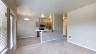 This single-floor residence offers a clean, contemporary design with an open-concept layout that connects the kitchen and living area in a cohesive and functional arrangement. The kitchen is outfitted with warm wood-toned cabinetry, a stainless steel microwave and range, and a peninsula countertop with an overhang that provides casual bar-style seating. A single-basin sink is integrated into the peninsula, and the kitchen benefits from flush-mount ceiling lighting that keeps the space bright and well-lit. A decorative pendant light hangs near the dining area, adding a refined touch to the overall aesthetic. The transition between the kitchen's hard-surface flooring and the living area's plush neutral carpet creates a clear visual distinction between the two zones while maintaining an open feel. The walls are finished in a soft gray tone, complementing the light-colored cabinetry and white countertops throughout. The living area is generously proportioned, offering ample room for furnishings. A hallway visible in the background leads to additional rooms, suggesting a well-organized floor plan. The overall ambiance is modern, clean, and move-in ready, with a neutral palette that provides a versatile foundation for personalization.
