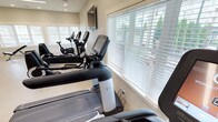 This fitness center is thoughtfully designed to support a comprehensive workout experience across multiple dedicated zones. The welcoming entry area features light hardwood-style flooring, soaring ceilings, and striking circular pendant light fixtures that create a bright, modern ambiance. A reception desk anchors the space, with colorful accent artwork and bold yellow wall accents adding energy to the interior.

The cardio area is well-appointed with a row of Life Fitness treadmills, ellipticals, and upright stationary bikes, all positioned near large windows fitted with white blinds that invite natural light while providing privacy. A wall-mounted flat-screen television serves the treadmill area for entertainment during workouts.

A separate strength and conditioning room offers additional equipment including an adjustable weight bench and stationary bike, surrounded on multiple sides by generous windows that frame views of the surrounding greenery, creating an airy, open feel underfoot with plush carpeted flooring.

The spacious studio or group fitness area showcases seamless light hardwood flooring, high ceilings, modern ring-style pendant lighting, and built-in shelving with a vibrant yellow accent wall — ideal for yoga, stretching, or group classes. Throughout the facility, recessed lighting and large windows ensure the space remains bright and inviting at all hours.