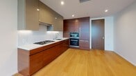 This contemporary single-floor residence showcases a thoughtfully designed open layout with a clean, modern aesthetic carried consistently throughout every space. Light hardwood flooring runs throughout, complementing the bright white walls and creating a warm yet refined atmosphere.

The kitchen is a standout feature, outfitted with rich walnut wood cabinetry paired with matte taupe upper cabinets, crisp white countertops, a white marble-style backsplash, and stainless steel appliances including a gas cooktop and double wall ovens. Under-cabinet lighting enhances the workspace, while recessed ceiling lights illuminate the open-plan area that flows seamlessly from the kitchen into the living space.

The living and bedroom areas benefit significantly from expansive floor-to-ceiling windows that frame sweeping panoramic city views stretching across the urban skyline to distant hills. Natural light floods these rooms, reinforcing the airy, elevated ambiance characteristic of a high-rise setting.

The bathroom is finished to a high standard, featuring full marble-look tile covering the floors, walls, and surround of the soaking tub. A glass-enclosed tub and shower combination, a wall-mounted toilet, and recessed lighting complete the spa-like environment. Overall, the space exudes a polished, contemporary character suited to modern urban living.