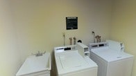 This shared laundry facility is a practical, well-organized space designed to serve the needs of its users efficiently. The room features multiple top-loading washing machines positioned along the wall, each with dedicated plumbing connections, making it easy to run several loads simultaneously. A utility sink is conveniently located nearby, providing a space for hand-washing or pre-treating garments before a full wash cycle. The walls are finished in a neutral, light tone that keeps the space feeling clean and open, while the layout ensures easy movement and access to all equipment. A posted "Laundry Room Rules" sign on the wall reflects a commitment to maintaining an orderly and respectful shared environment. Standard electrical outlets are accessible along the walls to accommodate additional appliances or equipment needs. The single-floor layout ensures straightforward accessibility, making this laundry room a reliable and no-frills communal resource. Overall, the space prioritizes function and cleanliness, offering residents or users a straightforward and well-maintained area to handle their laundry needs.