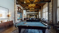 This single-story clubhouse in Spring, Texas, delivers an impressive blend of rustic and contemporary design throughout its thoughtfully arranged social spaces. The centerpiece of the main hall is a dramatic vaulted ceiling supported by exposed wood trusses and anchored by towering natural stone columns that rise from hardwood floors to create a grand, lodge-inspired atmosphere. Oversized floor-to-ceiling windows flood the space with natural light while framing outdoor views, and a full-size billiards table serves as a focal point beneath the soaring canopy.

The lounge area features rich leather sofas arranged around a wood-topped coffee table, complemented by patterned area rugs that add warmth and texture. Adjacent to the main lounge, a dining and entertainment zone offers a long communal table beneath industrial-style pendant lights, with stacked-stone accent walls and multiple wall-mounted televisions. A dedicated wet bar area includes dark cabinetry, a stone-tile backsplash, a built-in sink, and under-counter refrigeration—ideal for entertaining.

A separate media lounge provides a more intimate setting, with plush seating, a large flat-screen television, a beverage refrigerator, and moody, sophisticated décor. Together, these areas create a versatile and inviting community gathering space.